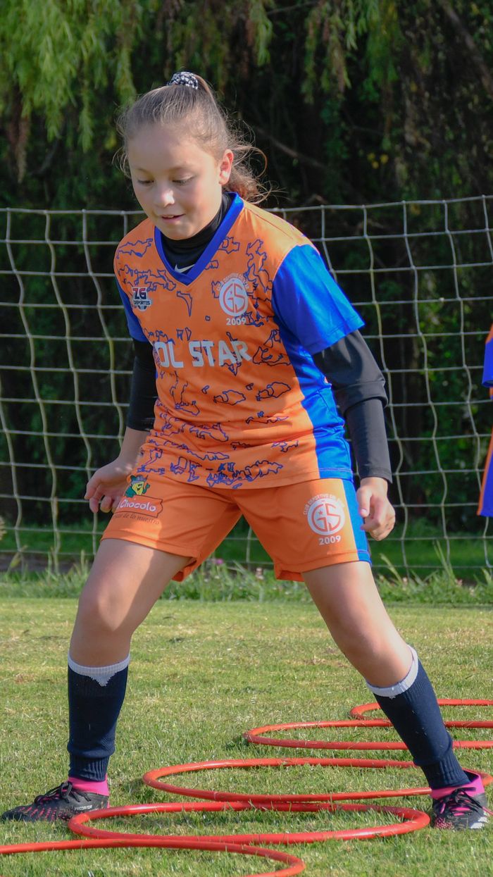 A young girl in an orange and blue kit practices soccer drills with agility rings on grass.